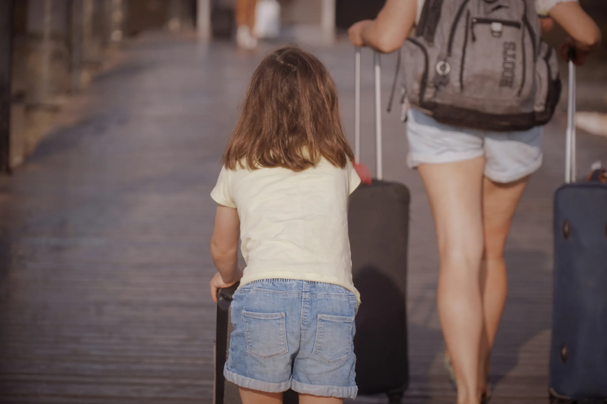 Child walking with a suitcase beside an adult while traveling for a family vacation.
